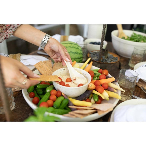 A close-up lifestyle shot of a hand dipping a cracker into a bowl of creamy hummus, surrounded by fresh vegetables like cherry tomatoes, cucumbers, carrots, and purple onions on a wooden table.