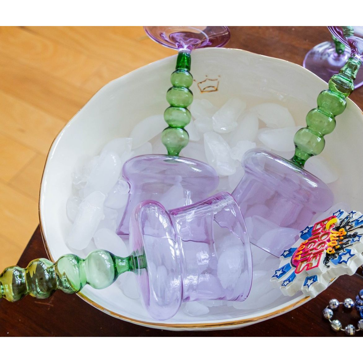 Three purple glasses with green stems rest upside down on ice in the Abigails Gold Rim Crown Serving Bowl.