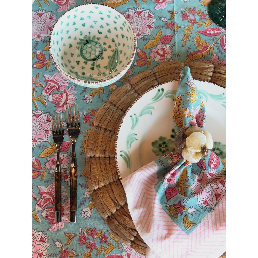 A styled flat-lay of a table setting featuring a turquoise floral tablecloth, a matching bowl, a large plate with a woven placemat, a pink striped napkin, and a floral napkin with a cotton flower accent.