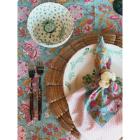 A styled flat-lay of a table setting featuring a turquoise floral tablecloth, a matching bowl, a large plate with a woven placemat, a pink striped napkin, and a floral napkin with a cotton flower accent.