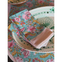 A close-up of a table setting featuring a turquoise floral placemat and matching napkins, with a folded pink striped napkin on a white patterned plate.