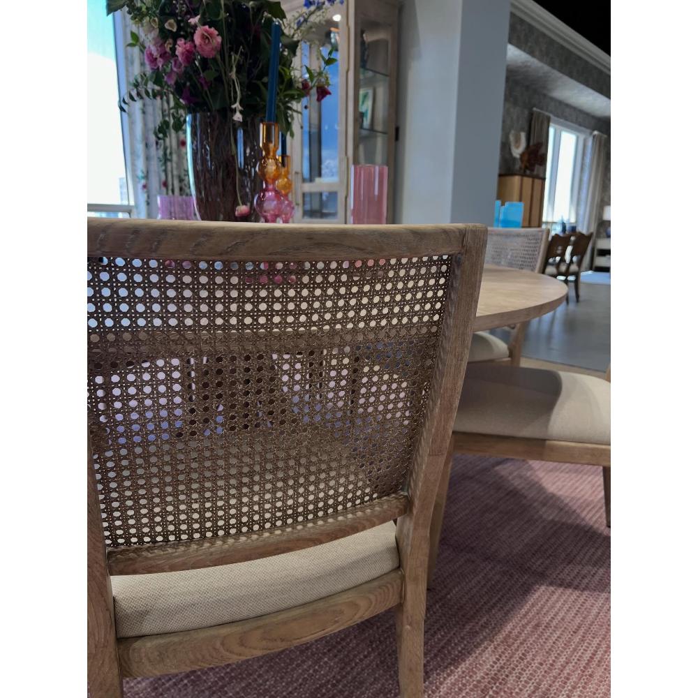 A close-up of a wooden dining chair with a woven cane backrest and a light beige upholstered seat cushion, shown in a dining room setting.