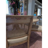 A close-up of a wooden dining chair with a woven cane backrest and a light beige upholstered seat cushion, shown in a dining room setting.