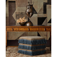 A styled interior scene featuring a wooden console table with drawers, a bronze abstract sculpture, a bowl of dried flowers, and a fringed blue-and-beige upholstered ottoman in front. The background is a geometric patterned wall.