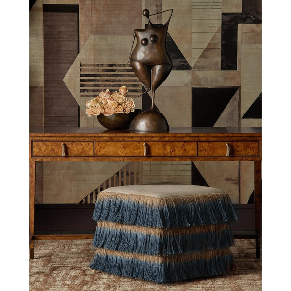 A styled interior scene featuring a wooden console table with drawers, a bronze abstract sculpture, a bowl of dried flowers, and a fringed blue-and-beige upholstered ottoman in front. The background is a geometric patterned wall.