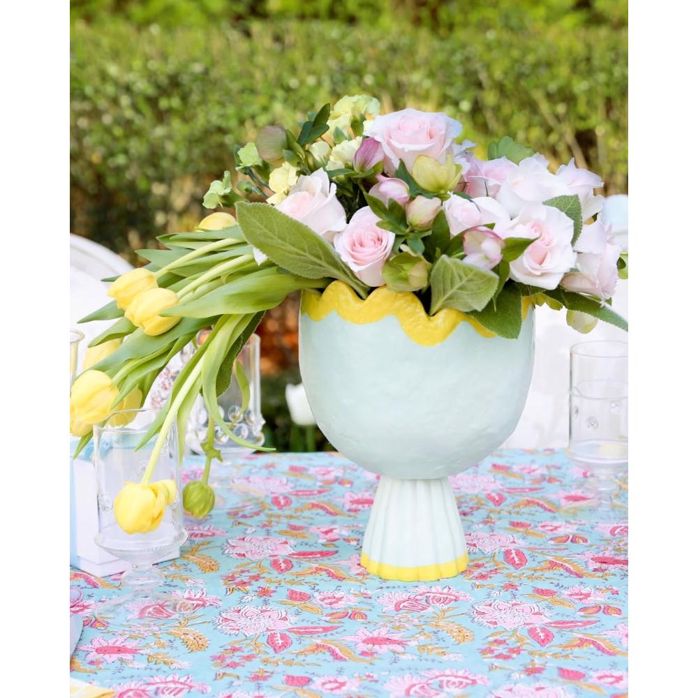 A pastel blue pedestal vase with a yellow scalloped rim, filled with pink and white roses and yellow tulips, set on a blue floral tablecloth outdoors.