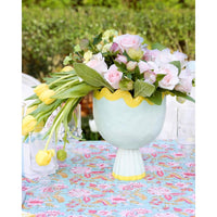 A pastel blue pedestal vase with a yellow scalloped rim, filled with pink and white roses and yellow tulips, set on a blue floral tablecloth outdoors.