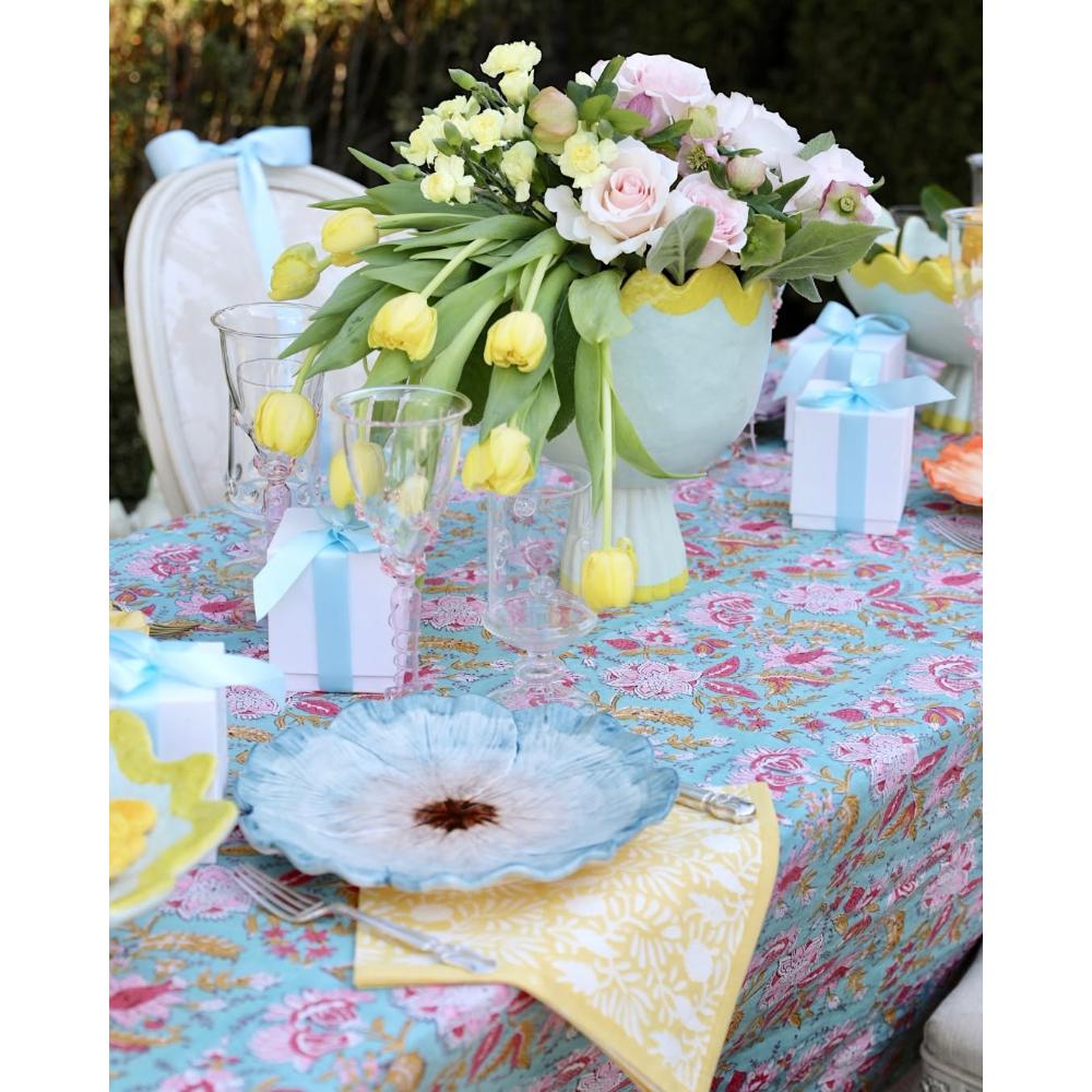 A styled outdoor table setting with a floral centerpiece of pink and yellow flowers in a light blue vase, a turquoise floral tablecloth, blue and yellow plates, yellow napkins, and small wrapped gift boxes tied with blue ribbons.