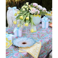 A styled outdoor table setting with a floral centerpiece of pink and yellow flowers in a light blue vase, a turquoise floral tablecloth, blue and yellow plates, yellow napkins, and small wrapped gift boxes tied with blue ribbons.