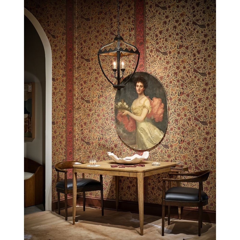 A vintage-style dining room scene featuring a wooden table with two dark chairs, a hanging lantern light, and a large oval portrait painting on a patterned wallpapered wall.