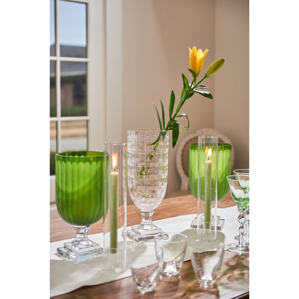 A styled dining table setting featuring green glass goblets, clear textured glass vases, lit taper candles, and a single yellow flower in a clear glass vase, set against a warm neutral background.