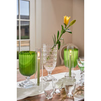 A styled dining table setting featuring green glass goblets, clear textured glass vases, lit taper candles, and a single yellow flower in a clear glass vase, set against a warm neutral background.