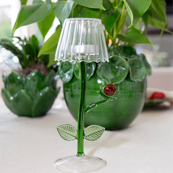 Decorative tealight glass lamp with green stem and leaf design on a surface with plants in the background.