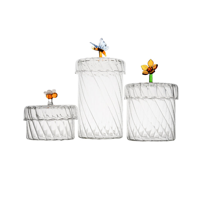 Three clear ribbed glass jars with matching lids, arranged by size on a white background. Each lid is topped with a small decorative floral or insect ornament in orange, yellow, green, and blue glass.