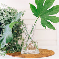 Le Sack Large Glass Vase by Abigails with big green leaves on a woven mat against a leafy backdrop on a white surface.