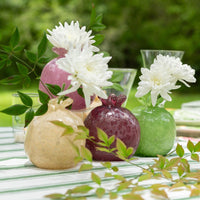 Three pomegranate-shaped glass vase toppers in beige, dark burgundy, and light green, styled with white flowers on a striped tablecloth in an outdoor garden setting.
