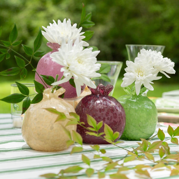 Three pomegranate-shaped glass vase toppers in beige, dark burgundy, and light green, styled with white flowers on a striped tablecloth in an outdoor garden setting.