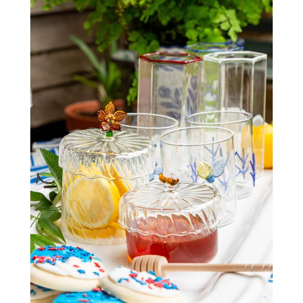 A set of clear glass canisters and tumblers with blue botanical patterns, styled on a white tablecloth with cookies, lemonade, and greenery in the background.
