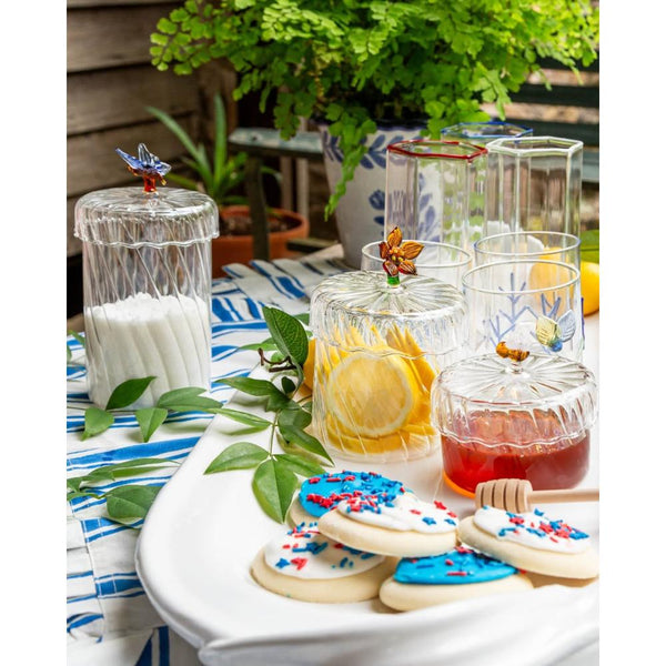 A styled tabletop scene with clear glass lidded jars and tumblers, some holding white sugar, lemon slices, and red beverage, alongside decorated sugar cookies on a white platter. The setting includes greenery, a blue-and-white striped cloth, and a wooden fence in the background.
