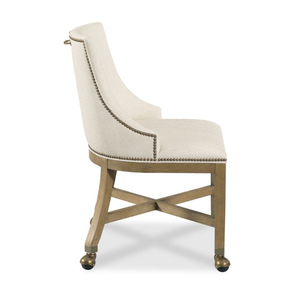 An upholstered accent chair with a curved cream fabric seat and back, detailed with nailhead trim, on a light wood frame with brass caster wheels.