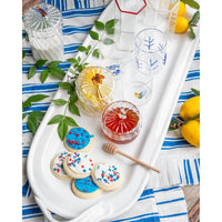 A styled tabletop scene with a white serving tray holding several clear glass tumblers with blue leaf motifs, a jar of red honey, a lemonade glass with lemon slices, cookies with red and blue sprinkles, a wooden honey dipper, and green leaves on a blue-and-white striped cloth with lemons nearby.