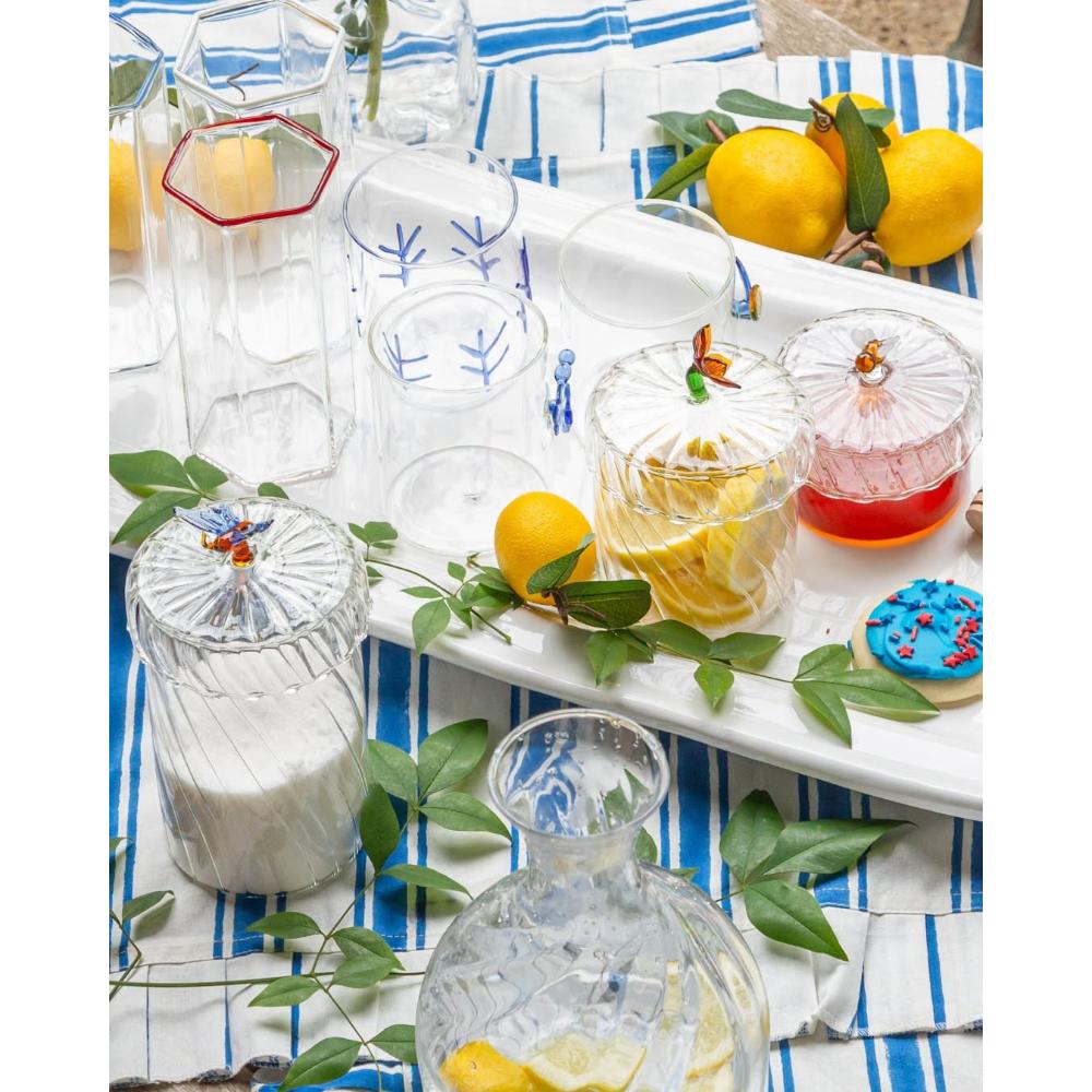 A styled tabletop scene with a white serving tray holding several clear glass tumblers with blue leaf motifs, a jar of red honey, a lemonade glass with lemon slices, cookies with red and blue sprinkles, a wooden honey dipper, and green leaves on a blue-and-white striped cloth with lemons nearby.