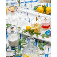 A styled tabletop scene with a white serving tray holding several clear glass tumblers with blue leaf motifs, a jar of red honey, a lemonade glass with lemon slices, cookies with red and blue sprinkles, a wooden honey dipper, and green leaves on a blue-and-white striped cloth with lemons nearby.