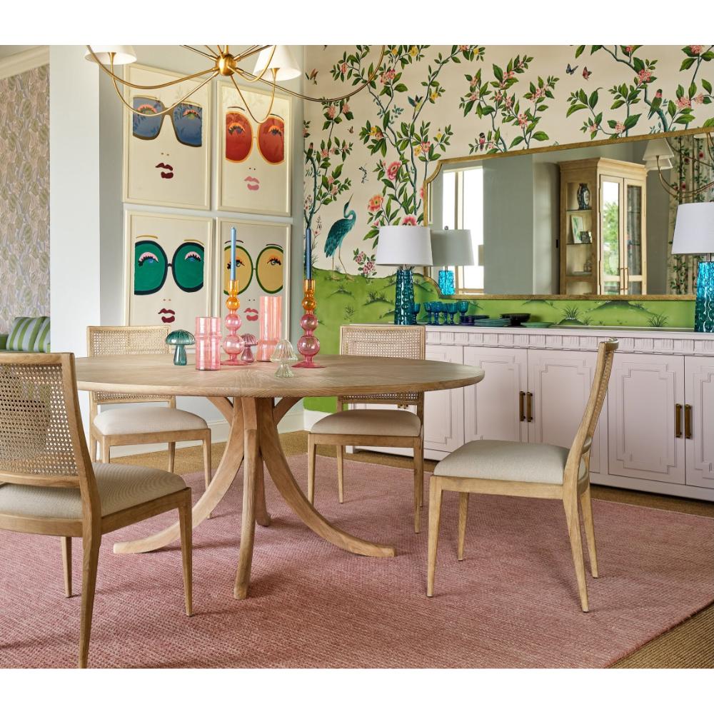 A mid-century modern round dining table with a natural wood pedestal base and four matching woven-back chairs with light beige upholstered seats, set on a dusty pink rug in a decorated dining room.