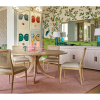 A mid-century modern round dining table with a natural wood pedestal base and four matching woven-back chairs with light beige upholstered seats, set on a dusty pink rug in a decorated dining room.