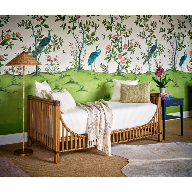 A bamboo daybed with white bedding and pillows in a styled bedroom, set against a distinctive wallpaper featuring peacocks, birds, and flowers on a green and white background.