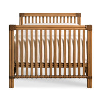A wooden crib with vertical slats and rounded corner brackets, shown from the front on a clean white background. A white mattress is visible inside.