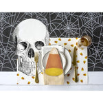 Create the perfect spooky tablescape for your Halloween party with a Halloween-themed table setting featuring a large skull print, candy corn decorations, and utensils placed on a Hester & Cook Halloween Spiderweb Paper Table Runner.
