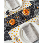 A spooky tablescape featuring the Hester & Cook Halloween Spiderweb Paper Table Runner, candy corn, a small pumpkin, and gray napkins on plates brings the perfect eerie charm to your Halloween party.
