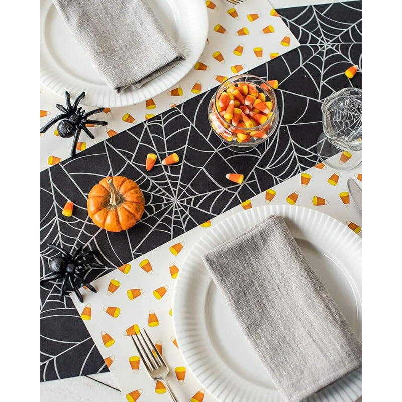 A spooky tablescape featuring the Hester & Cook Halloween Spiderweb Paper Table Runner, candy corn, a small pumpkin, and gray napkins on plates brings the perfect eerie charm to your Halloween party.