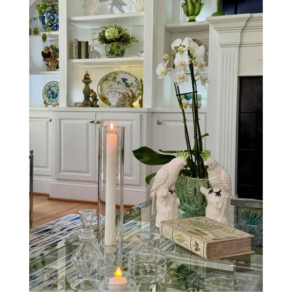A lit taper candle inside an Abigails Candlestick Holder with Hurricane Cylinder on a glass coffee table.