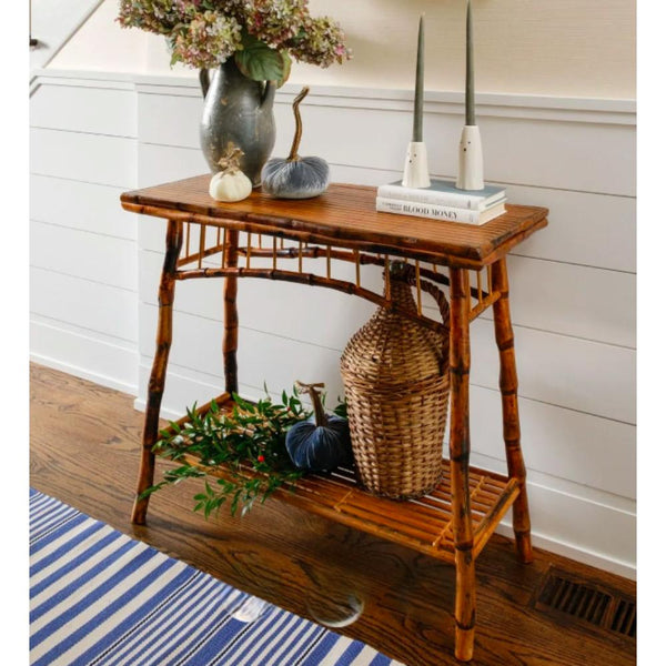 A rustic bamboo console table styled with a gray vase of flowers, pumpkins, candles, and a woven basket on a wooden floor against a white paneled wall.