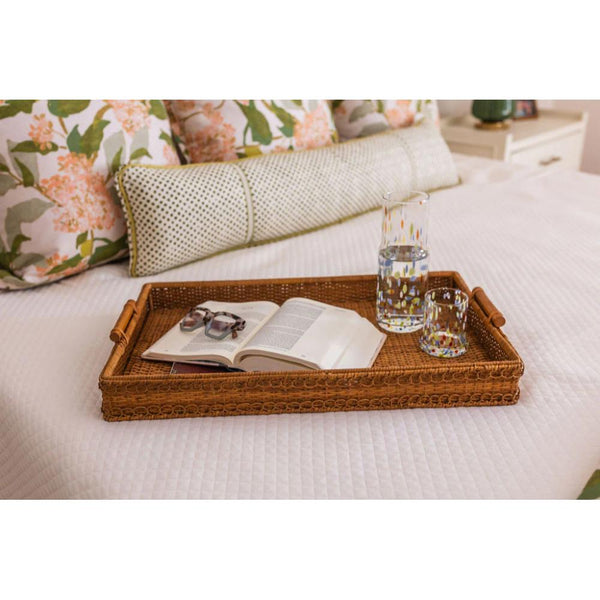 Rattan tray with book, glasses, and water bottle on a bed with floral pillows.