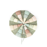 A round patchwork christmas tree skirt with a sage green base and multiple stitched fabric segments in pastel pink, beige, mint green, and light blue, finished with a drawstring closure, shown on a plain white background.