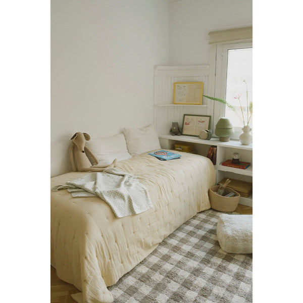 A cozy, minimalist bedroom scene featuring a bed dressed in a textured cream quilt and matching pillow shams, styled with a light green knit throw blanket and a plush dog toy. The room has white walls, built-in white shelving with decorative items, a window, and a beige-and-white gingham checkered rug.