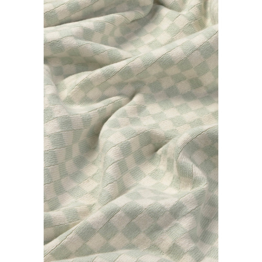 A close-up of a soft, textured fabric with a light sage green and off-white checkerboard pattern, shown with gentle folds.