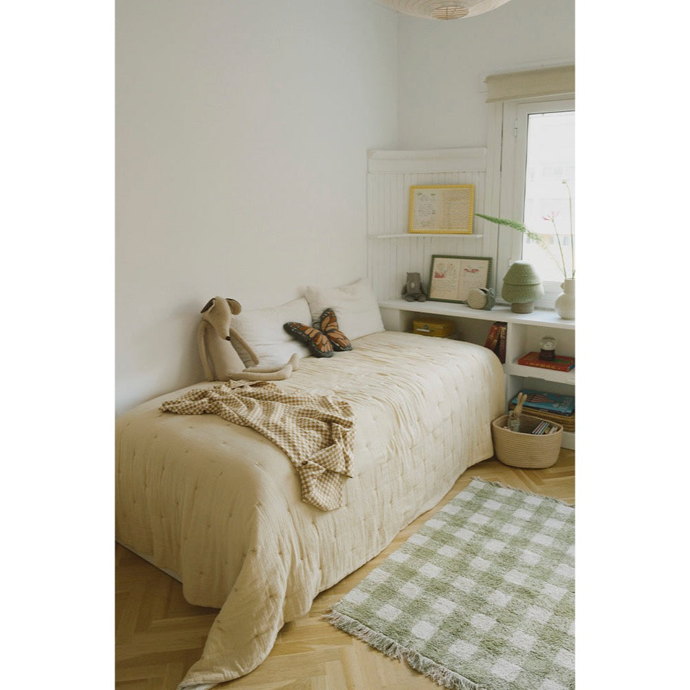 A cozy single bed dressed in a cream quilted coverlet and matching knit throw, styled in a bright, minimalist bedroom with white walls, a window, shelving, and a green-and-white checkered rug on the wood floor.