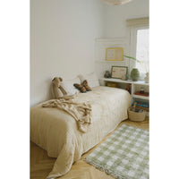 A cozy single bed dressed in a cream quilted coverlet and matching knit throw, styled in a bright, minimalist bedroom with white walls, a window, shelving, and a green-and-white checkered rug on the wood floor.