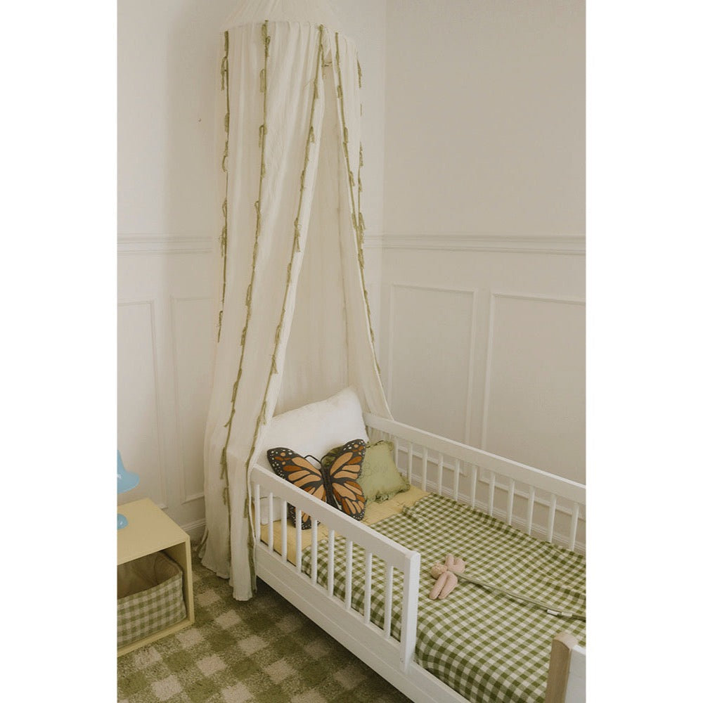 A white wooden toddler bed with a matching canopy tent draped over it, styled with a green and white gingham sheet, a butterfly pillow, and a small pink toy, set against a white paneled wall.