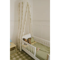 A white wooden toddler bed with a matching canopy tent draped over it, styled with a green and white gingham sheet, a butterfly pillow, and a small pink toy, set against a white paneled wall.