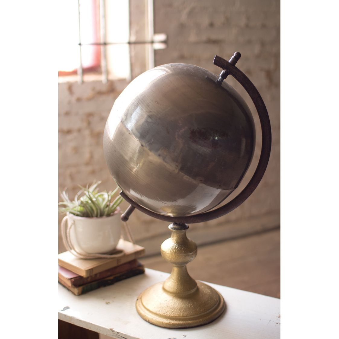 A BSEID Metal Gunmetal Gray + Rustic Brass Finish Globe sits by a plant and books on a white table near a window.
