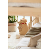 A beige knitted dog-shaped floor basket with a red collar sits on a light-colored rug in a cozy, softly lit room next to a potted plant.