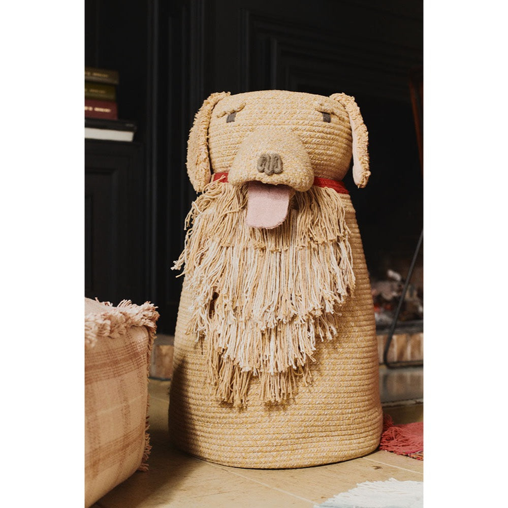 A woven straw dog-shaped storage basket with floppy ears, a pink tongue, and a long fringe beard, styled in a cozy home setting.