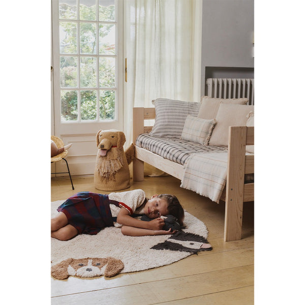 A child lies on a round cream rug with a dog face motif in a bright, modern children's bedroom. A light wood daybed with striped bedding, a dog-shaped stool, and a window are visible in the room.