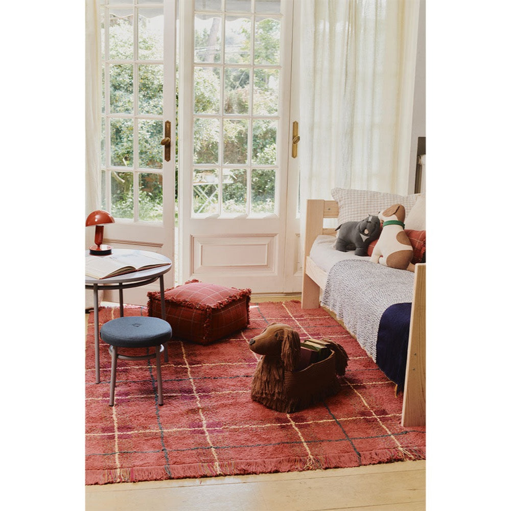 A styled interior room scene showing a red patterned area rug on a wooden floor, with a daybed, stool, side table, and a dog lying on the daybed. The product appears to be the rug, shown in a real home environment rather than a clean studio setup, so subject_type is Lifestyle.