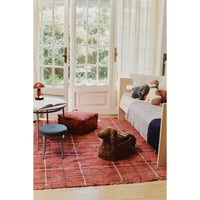 A styled interior room scene showing a red patterned area rug on a wooden floor, with a daybed, stool, side table, and a dog lying on the daybed. The product appears to be the rug, shown in a real home environment rather than a clean studio setup, so subject_type is Lifestyle.